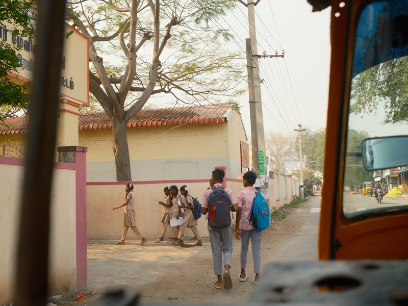 Children walking along a street with backpacks, framed by trees and buildings in a rural setting.