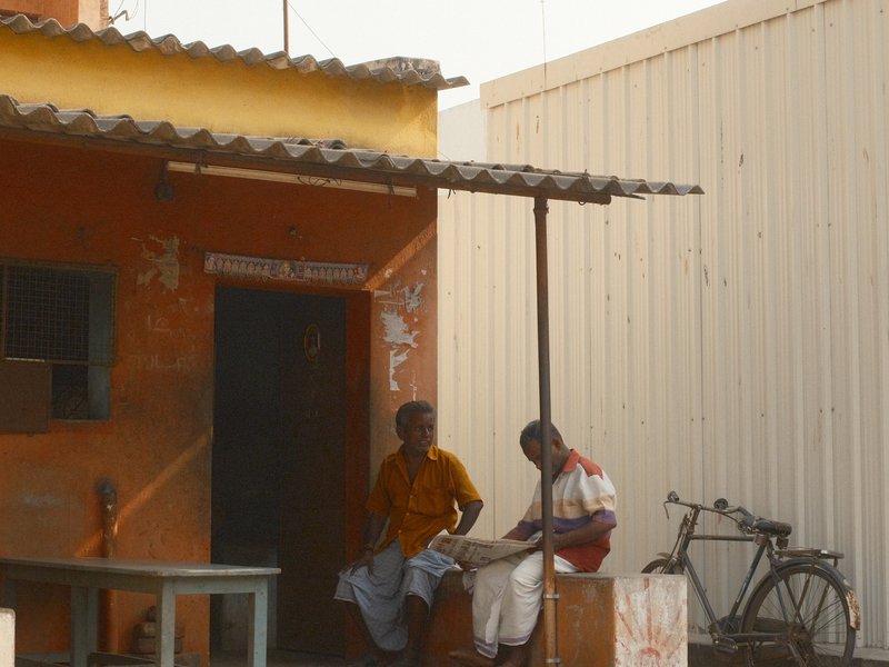 Two men sitting outside a rustic building with a bicycle nearby, under a shaded area.