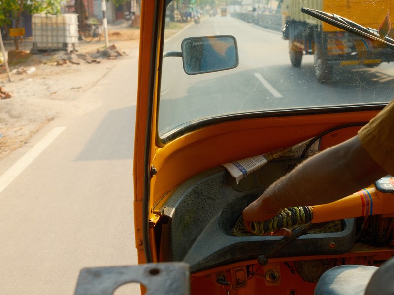 View from inside an auto rickshaw showing the driver's hand on the gear shift and a road ahead.