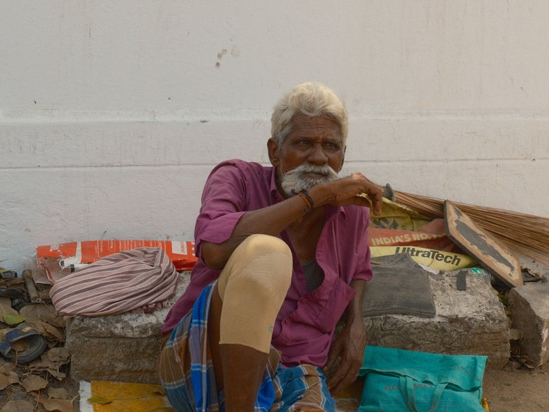 An elderly man sitting against a wall, wearing a purple shirt and holding a stick, with various items around him.