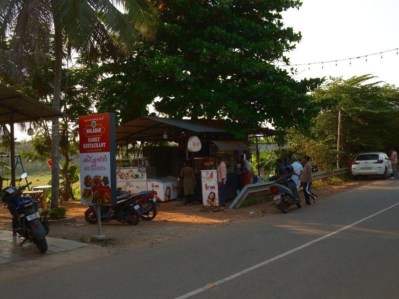 A roadside food stall with people, motorcycles, and a car under palm trees during golden hour.