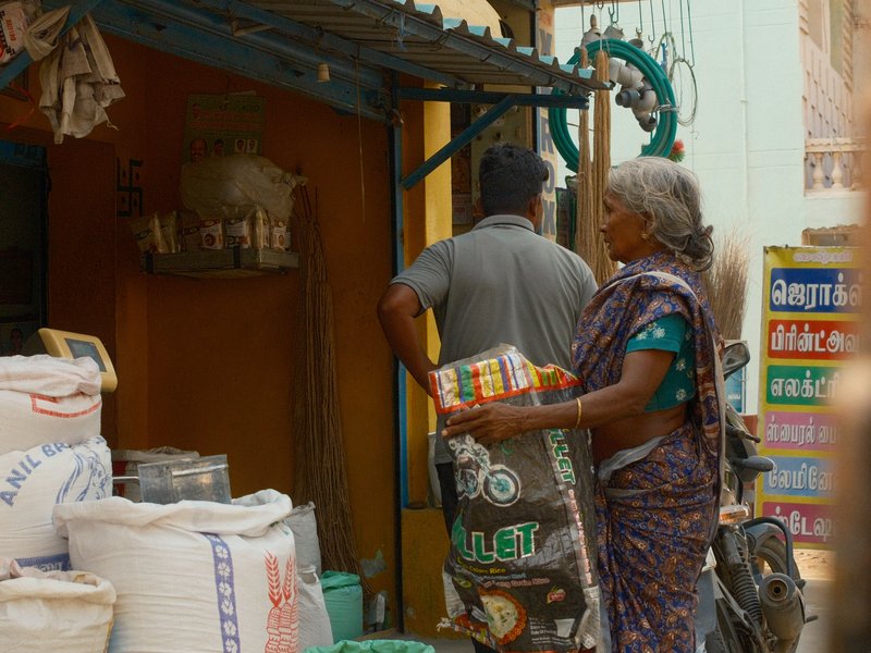 A woman carrying a bag of goods walks past a shop with sacks and a man standing nearby.