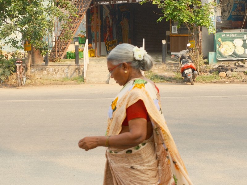 A woman in traditional attire walking on a street with a shop in the background.