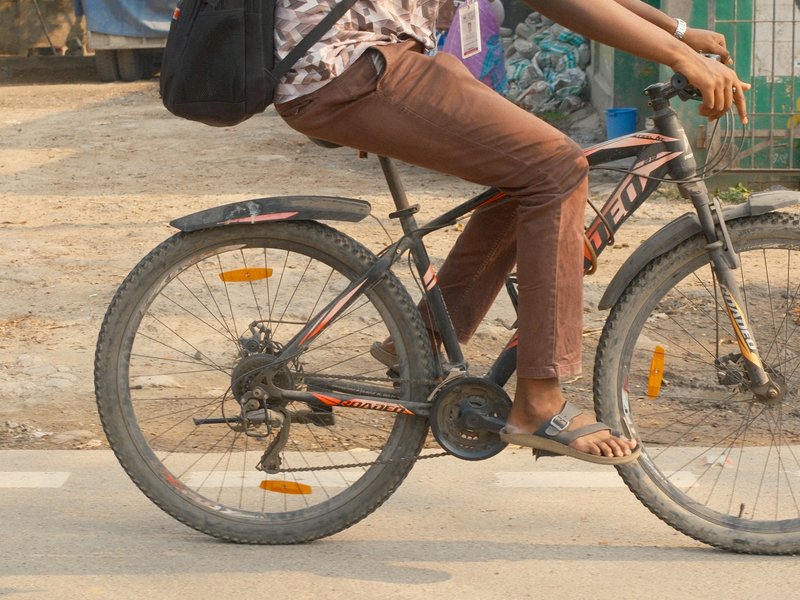 A person riding a bicycle on a dirt road, wearing brown pants and a patterned shirt.