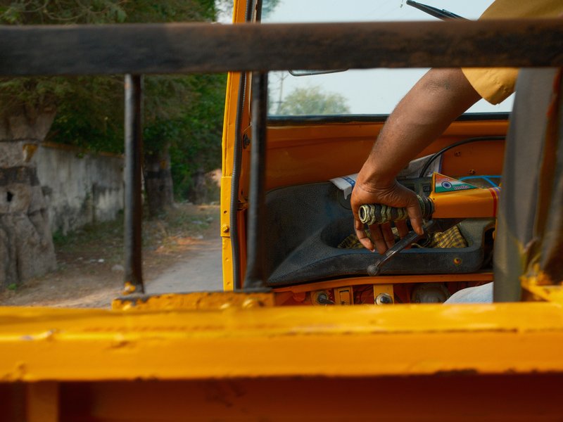 A close-up of a person's hand gripping the steering wheel of a yellow auto rickshaw on a rural road.