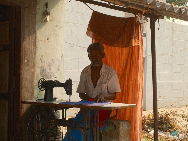 An elderly man sitting at a sewing machine under a shaded area with a curtain.