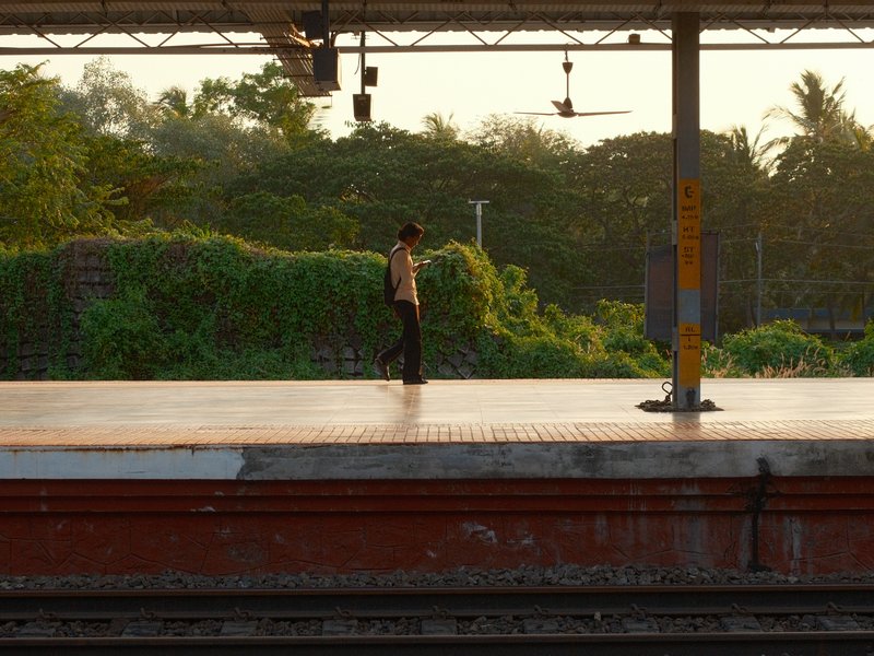 A solitary figure stands on a train platform surrounded by lush greenery at sunset.