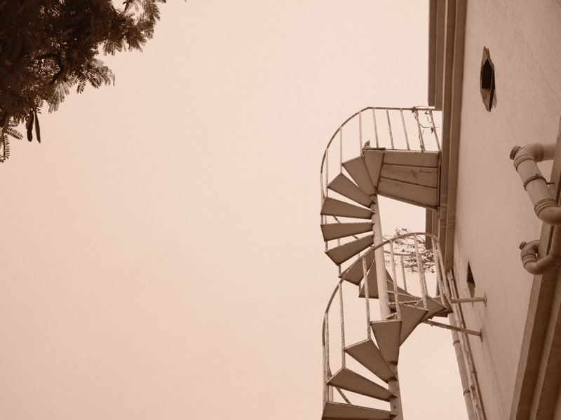 A spiral staircase ascending alongside a building, with a tree partially visible in the frame.