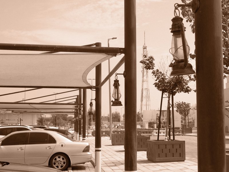 A sepia-toned view of a parking area with vintage lanterns, cars, and distant buildings.