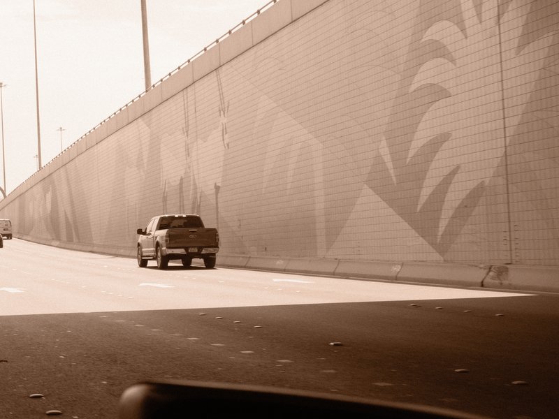 A pickup truck driving on a highway beside a mural-covered wall under a bright sky.