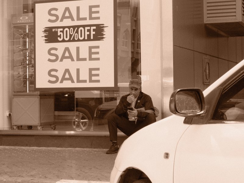 A man sitting outside a store with a sale sign, eating food, with a car in the foreground.