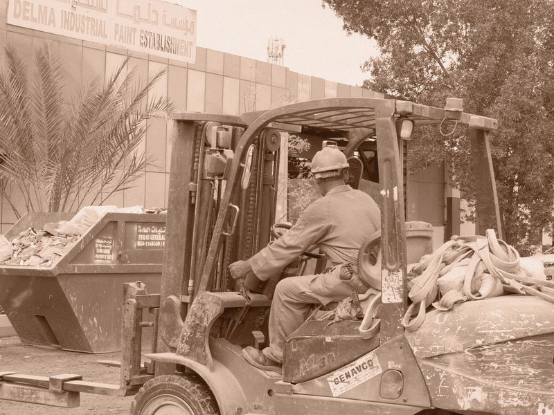 A worker operating a forklift in front of a building, surrounded by debris and greenery.