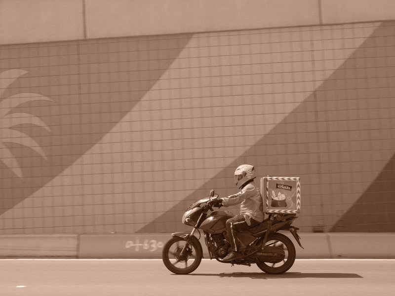 A motorcycle courier rides past a colorful mural with palm tree patterns on a city street.