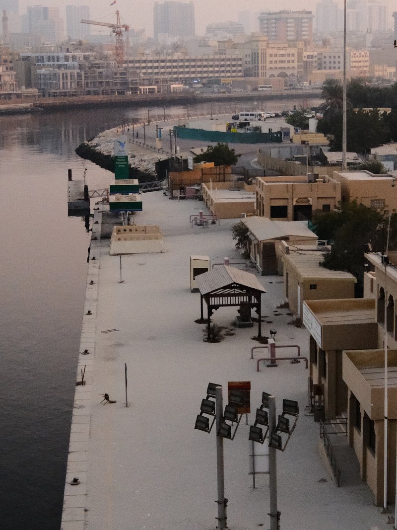A quiet waterfront promenade with buildings and lights along the water's edge during dusk.