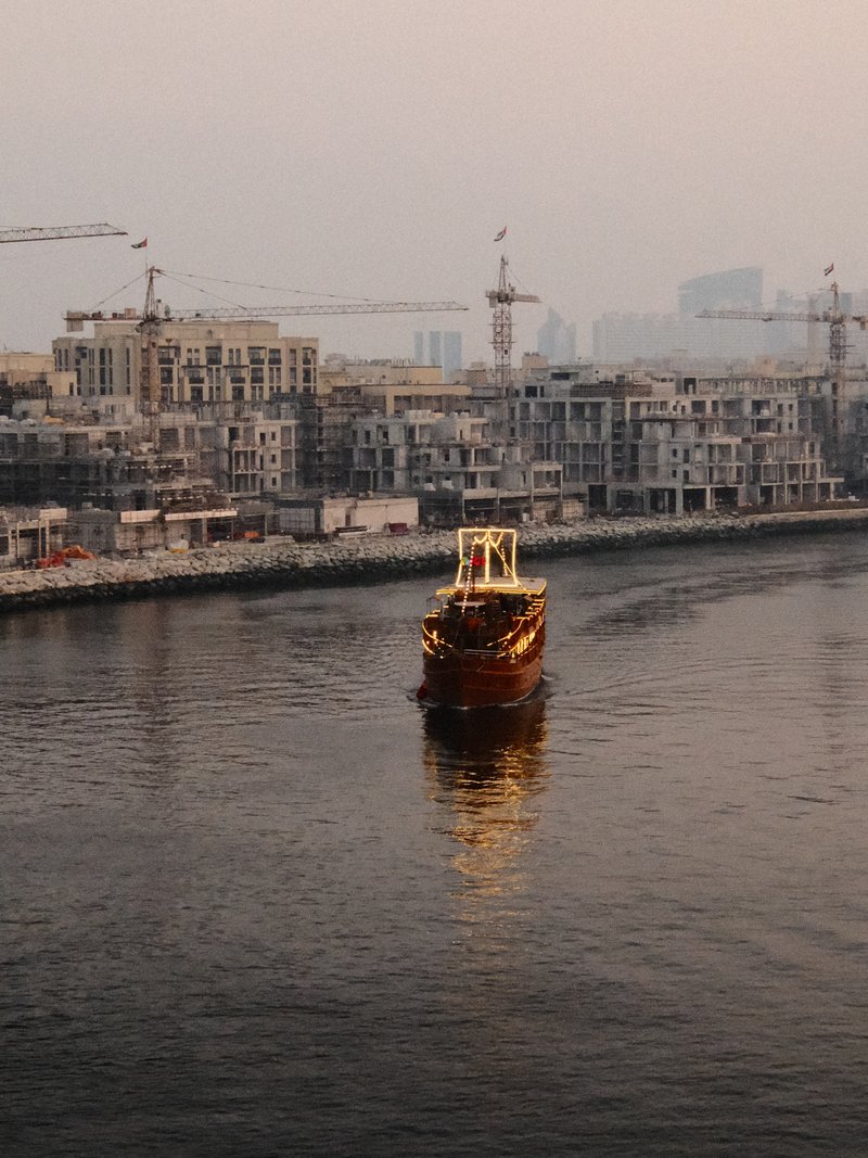 A boat navigating a calm river surrounded by construction sites and cranes in the background.
