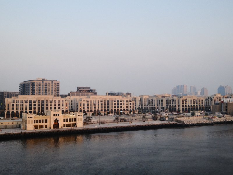 A waterfront view of modern buildings along a river at dusk, with soft light reflecting on the water.