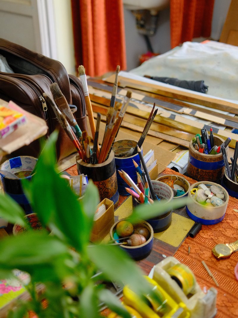 A cluttered artist's workspace with various tools, brushes, and plants in a cozy indoor setting.