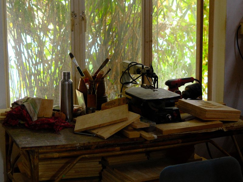 A cluttered wooden table with tools and materials, sunlight filtering through a window with greenery outside.