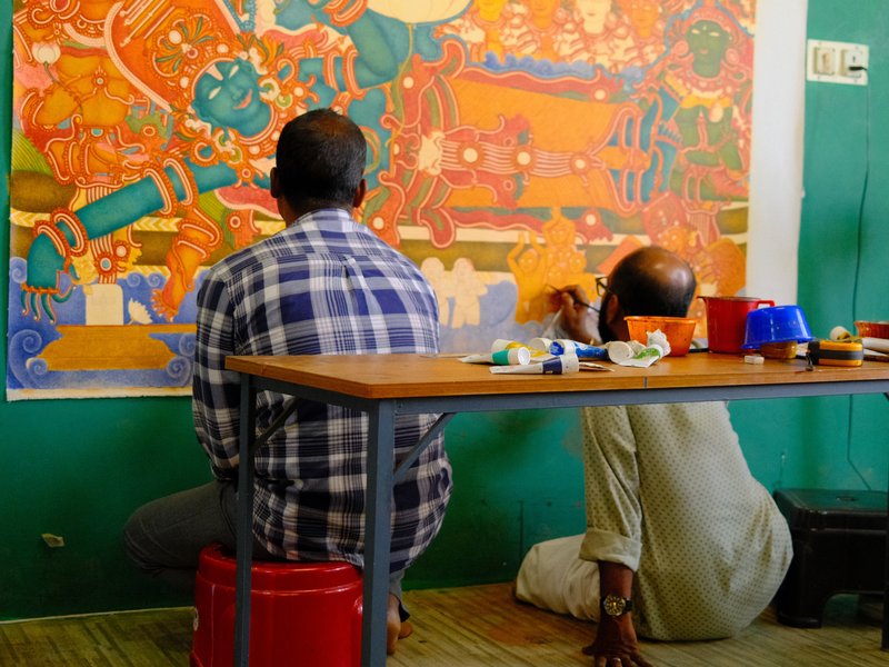 Two artists painting a colorful mural on a wall, seated at a table with art supplies.