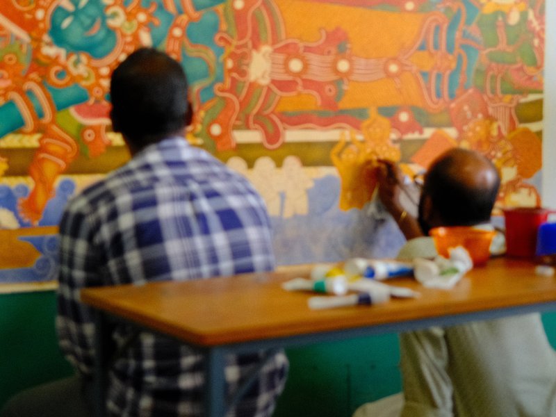 Two artists painting a colorful mural on a wall, seen from behind, with art supplies on a table.