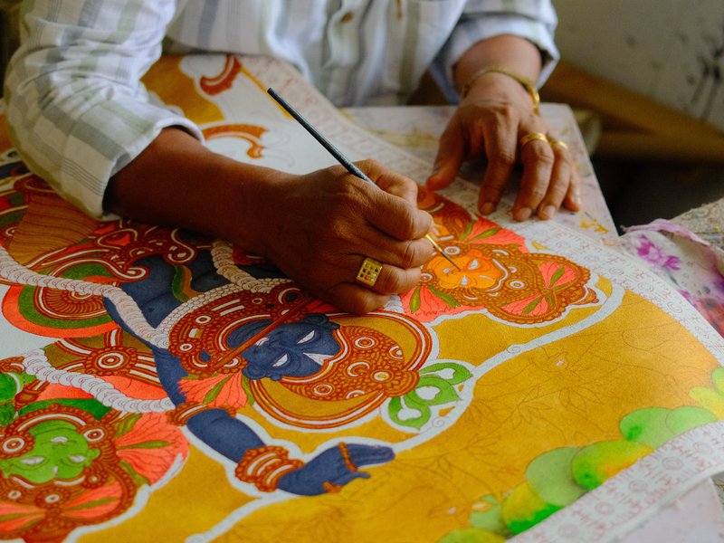 A close-up of a person's hand painting a colorful traditional artwork featuring deities.