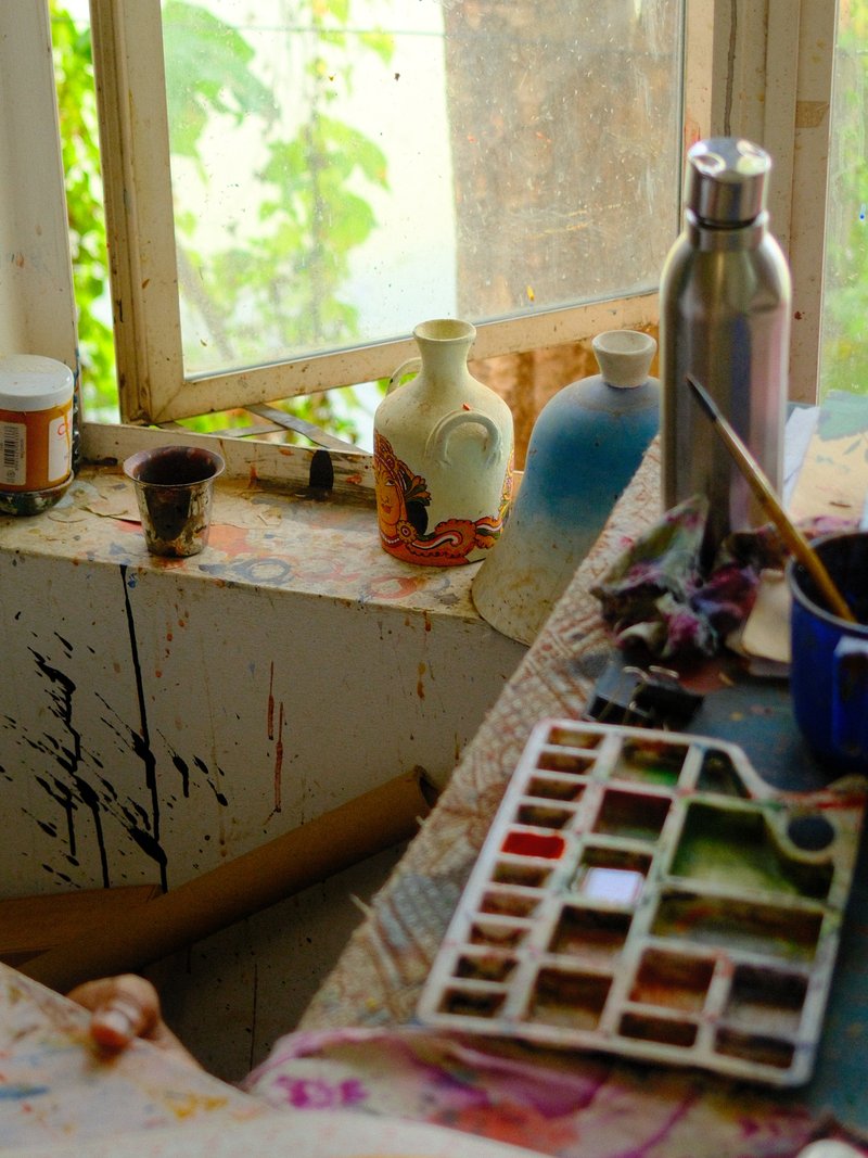 A cluttered artist's workspace with paint palettes, jars, and a window letting in natural light.