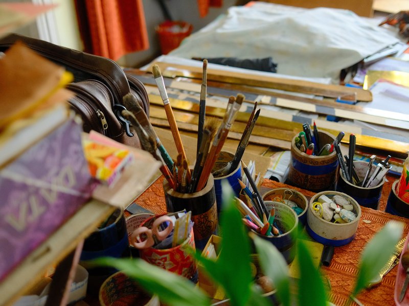 A cluttered artist's workspace with paintbrushes, paint pots, and various art supplies on a table.