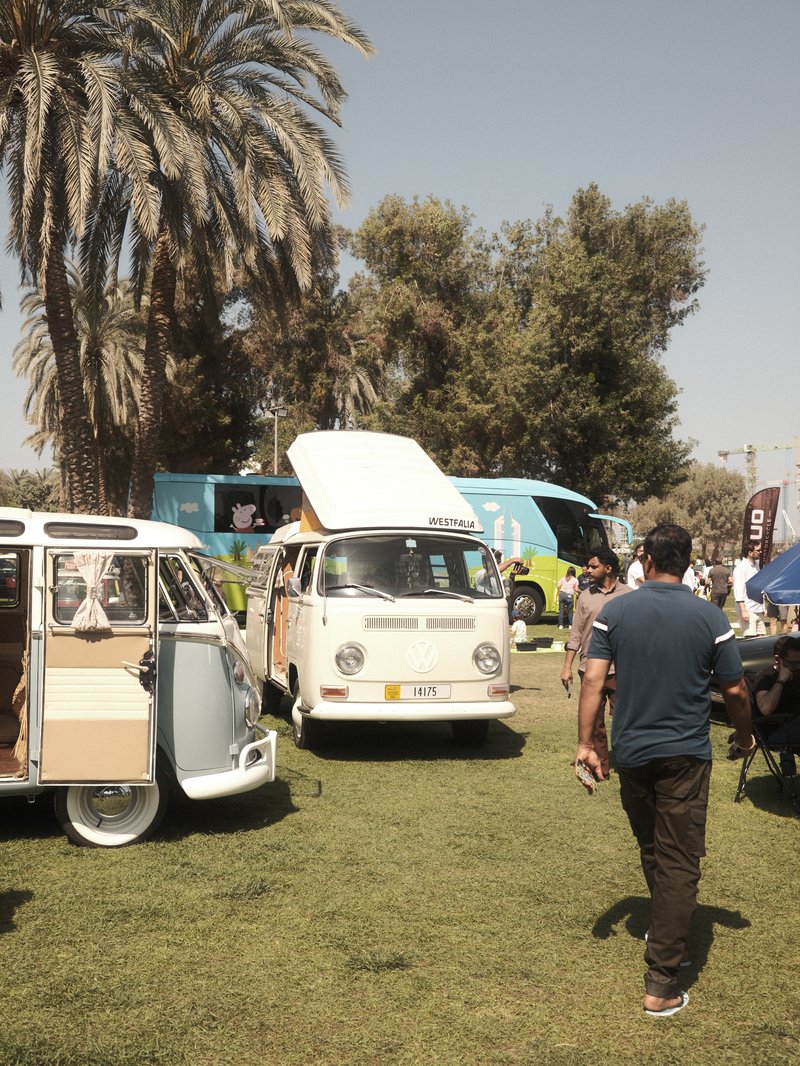 Two vintage vans parked on green grass with palm trees in the background and people walking around.