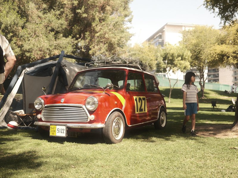 A vintage red car with racing stripes parked on green grass near a tent, with a child walking nearby.