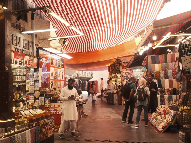 A vibrant market scene with colorful stalls and people browsing various goods under a striped canopy.