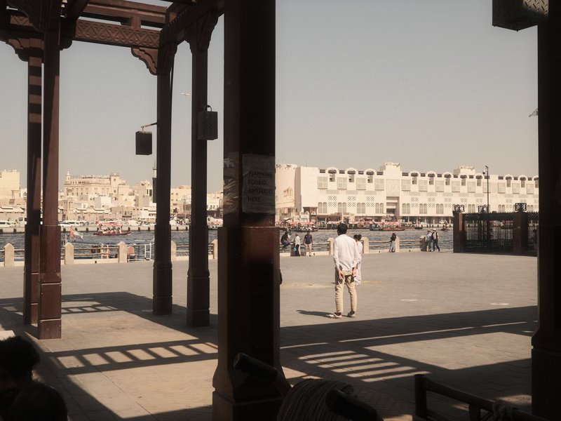 A lone figure stands under a wooden structure, overlooking a waterfront scene with buildings in the background.