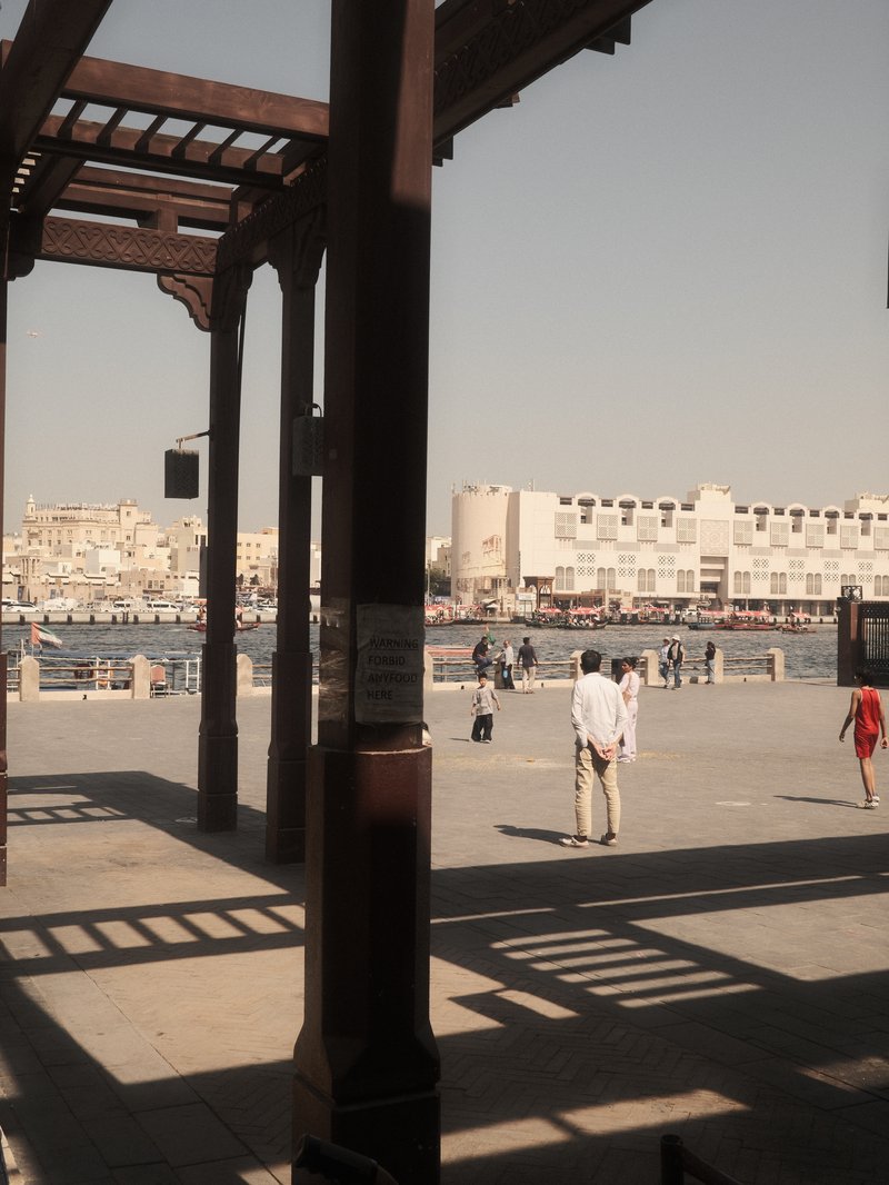 View of a waterfront promenade with people walking under wooden arches, buildings in the background.