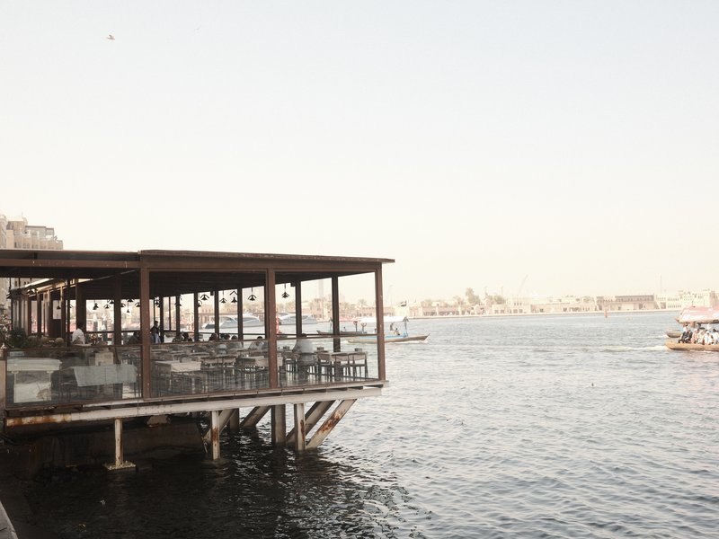 A waterfront restaurant with outdoor seating overlooking a calm river during the day.