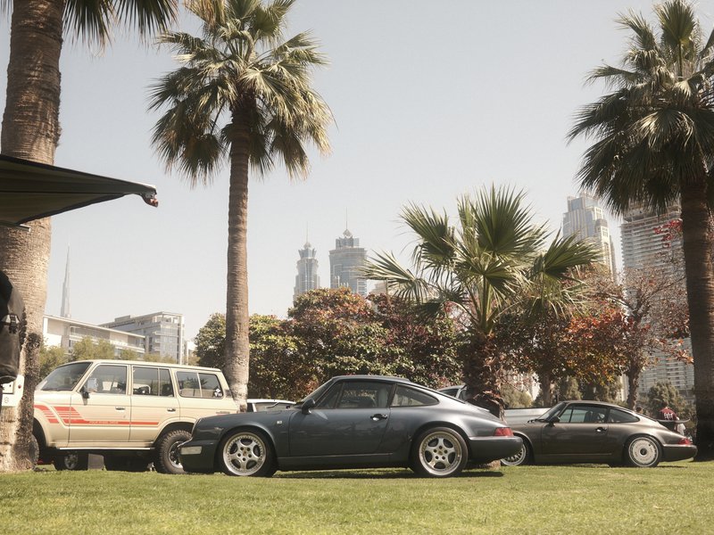 A vintage car parked on a grassy area with palm trees and city skyline in the background.
