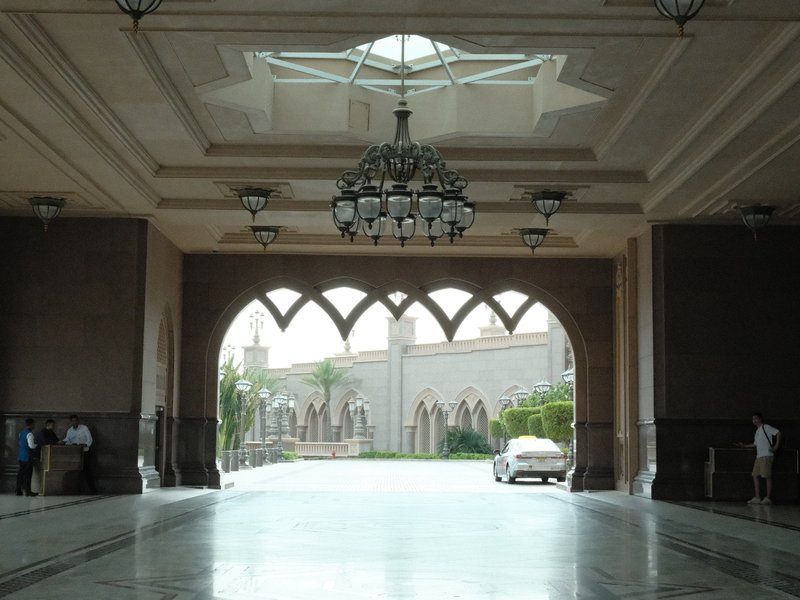 Interior view of a grand entrance hall with arches and a chandelier, leading to an outdoor area.