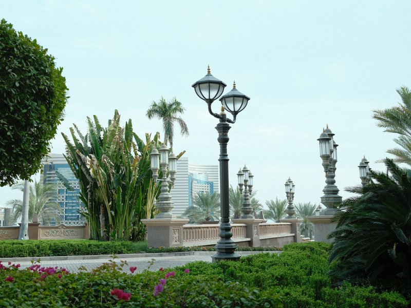 A scenic view of a park with decorative lampposts, palm trees, and lush greenery under a cloudy sky.