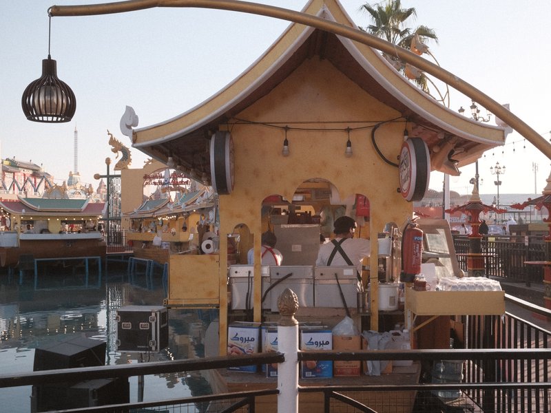 A colorful food stall with a unique architectural design by the water, surrounded by palm trees.