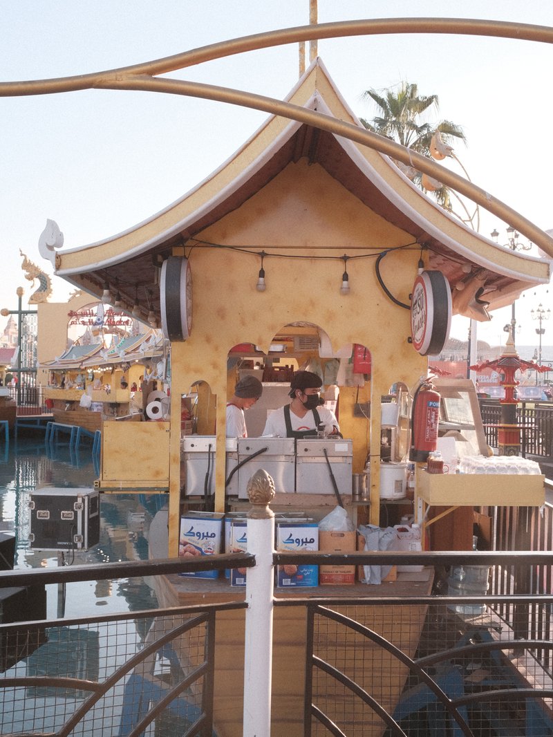 A whimsical yellow building resembling a boat, surrounded by water and palm trees in a carnival setting.