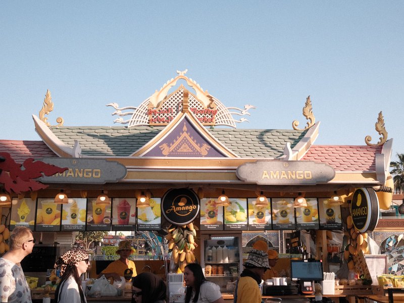 Colorful food stall with ornate roof and vibrant signage, bustling with people in a sunny outdoor setting.