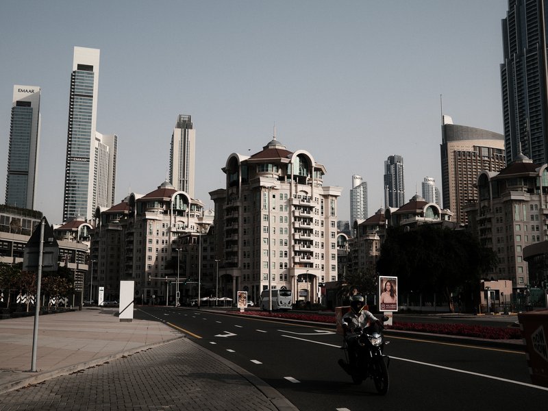 A motorcycle rider on a road with modern skyscrapers and unique architecture in the background.