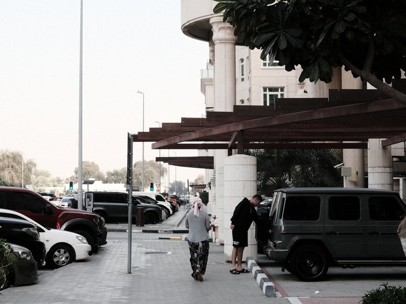A street scene with parked cars and pedestrians near a modern building under a clear sky.