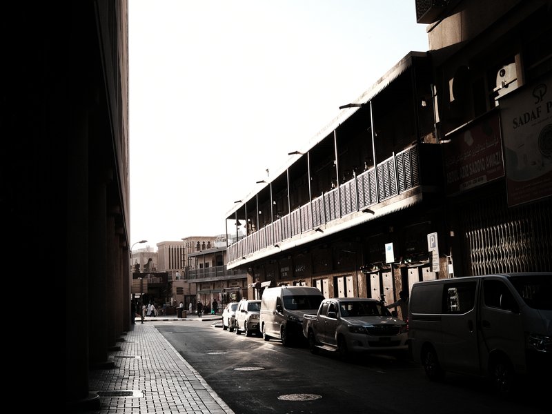 A narrow urban street with parked vehicles and buildings under bright sunlight, creating strong contrasts.