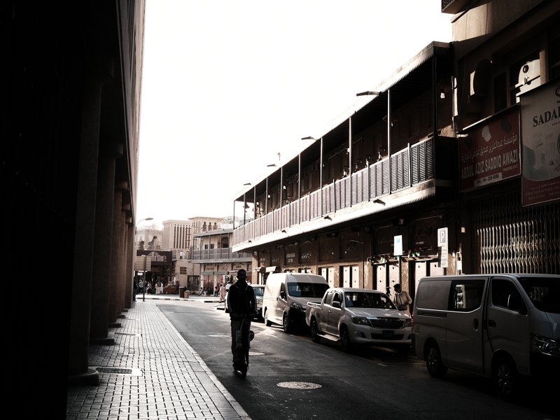 A lone figure walking down a narrow urban street with buildings and parked cars under soft light.