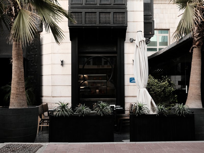 A modern restaurant entrance with palm trees and a white curtain, featuring a sleek black facade.