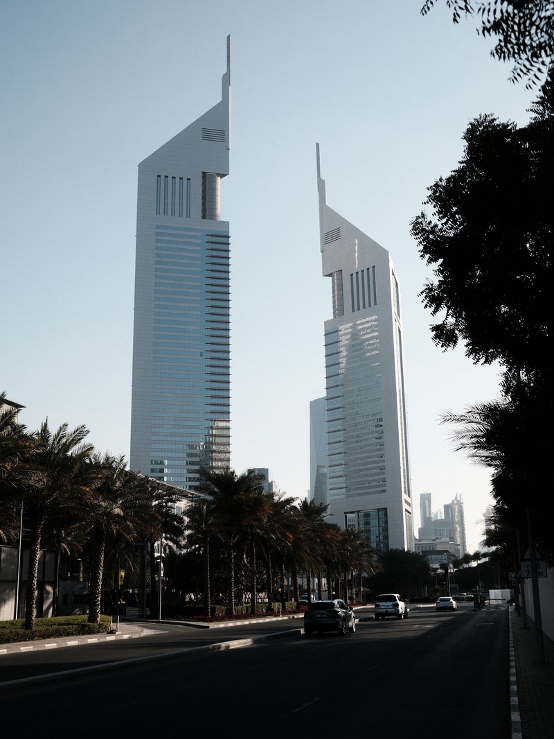 Two modern skyscrapers with unique architectural designs against a clear sky, lined by palm trees.