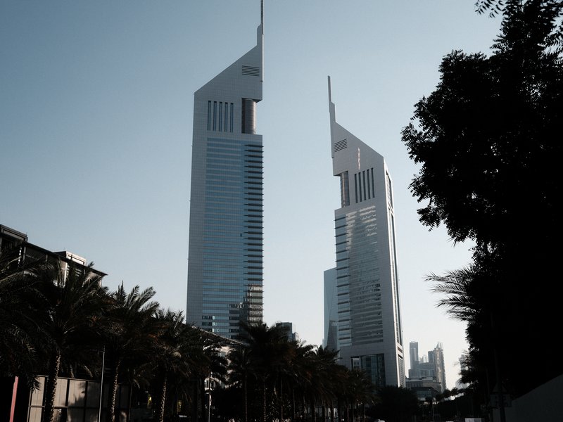 Two modern skyscrapers with pointed tops against a clear sky, surrounded by palm trees.