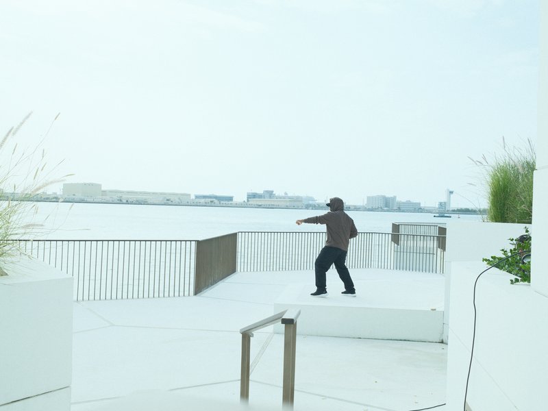 A person in a brown jacket stands on a white terrace by the water, with a hazy skyline in the background.