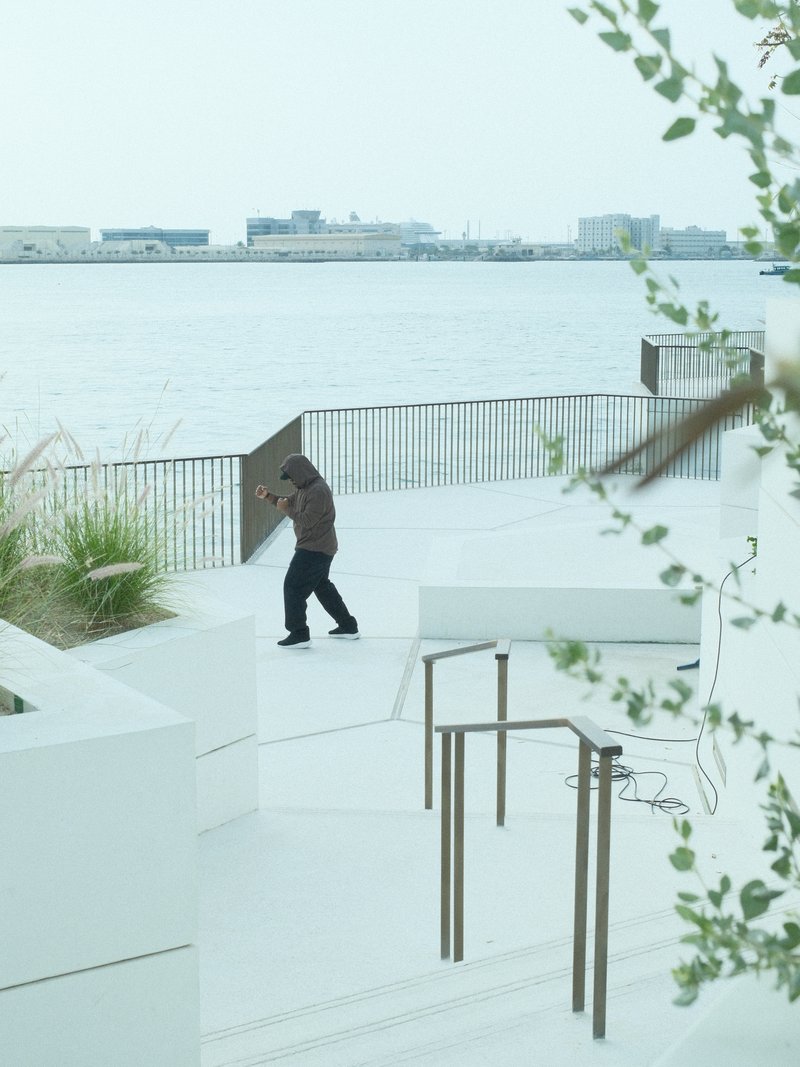 A person walking along a waterfront promenade with modern architecture and greenery.