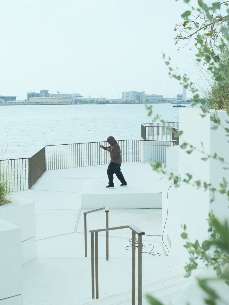 A person in a brown jacket stands on a white terrace overlooking a calm water body with distant buildings.