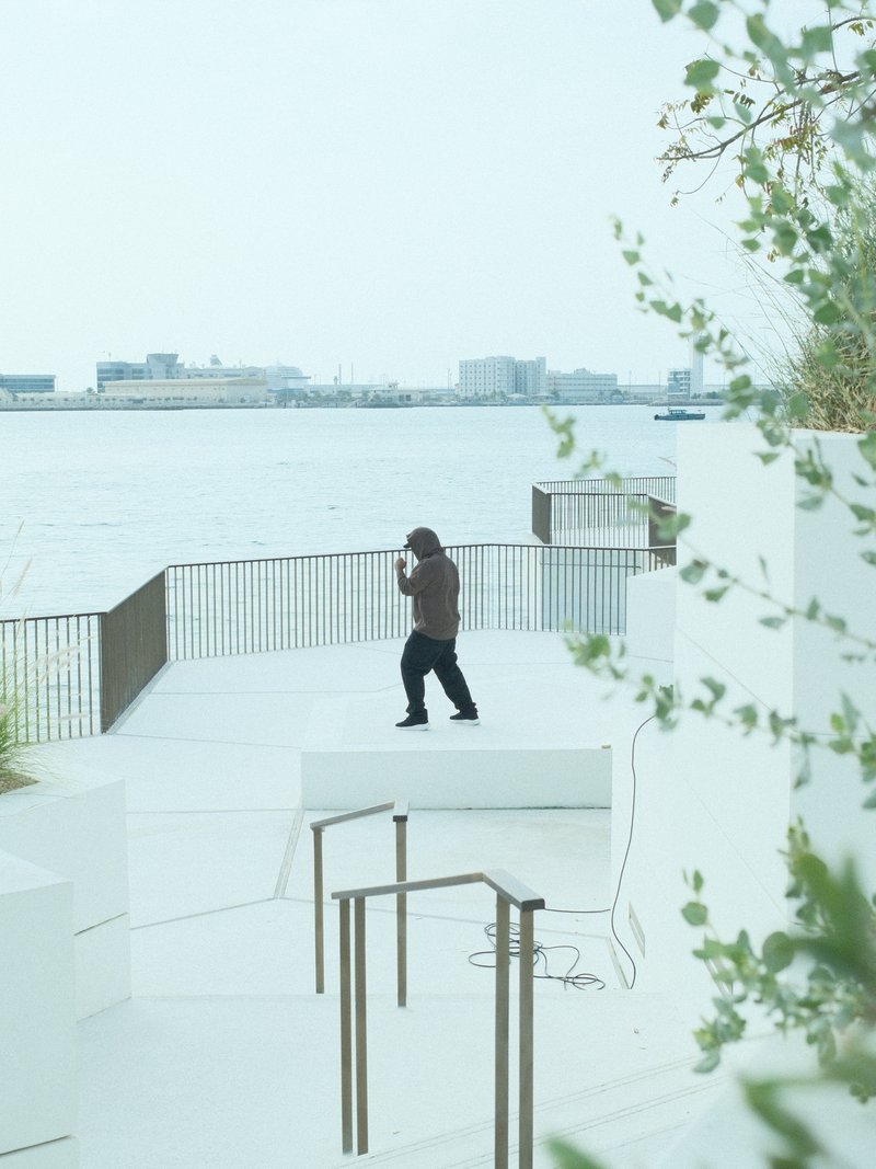 A person in a brown jacket stands on a white terrace overlooking a calm body of water.